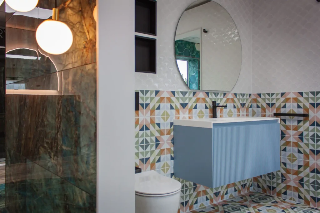 Modern bathroom with patterned tiles, large round mirror, blue floating vanity, and globe pendant lights. Reflective surfaces create a spacious feel.