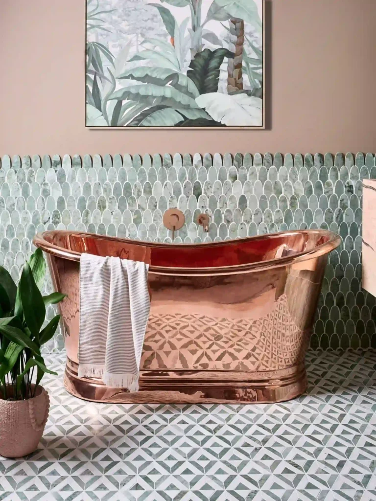 A luxurious bathroom with a copper bathtub, green-patterned tiles, tropical wall art, potted plant, and a striped towel draped over the tub.