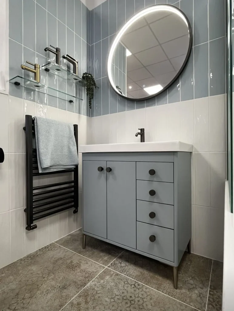 Modern bathroom with blue tiles, a round mirror, sleek black fixtures, and a light blue vanity. Towel on black heated rack.