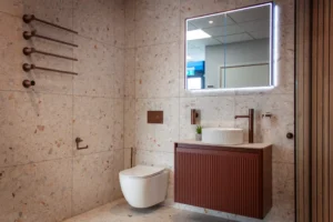 Modern bathroom with terrazzo tiles, wall-mounted toilet, vanity with round sink, mirror with LED lighting, towel bars, and a small potted plant.