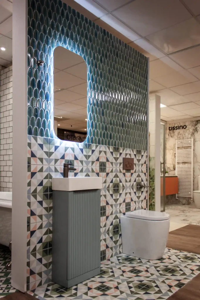 A modern bathroom features geometric patterned tiles, a backlit mirror, a small sink, and a toilet within a tiled showroom display setting.