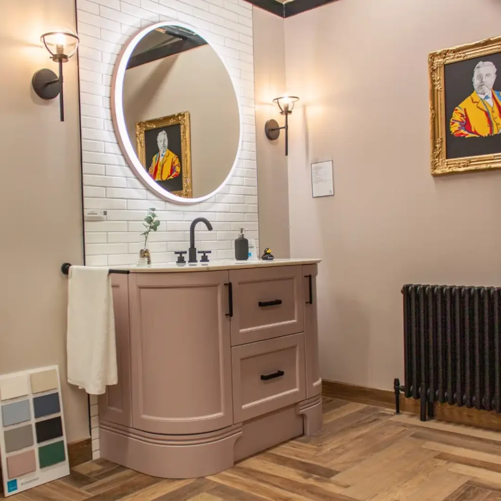 Modern bathroom with a lighted round mirror, pink vanity, framed artwork, and wooden floor. Stylish lamps and color samples on display.