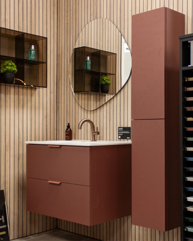 A modern bathroom setup with a circular mirror, maroon floating vanity, wall shelves with plants, and an assortment of color samples nearby.