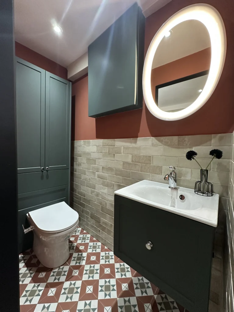 Modern bathroom with geometric floor tiles, dark cabinetry, circular illuminated mirror, maroon walls, and decorative flowers on white sink. Bright, stylish atmosphere.