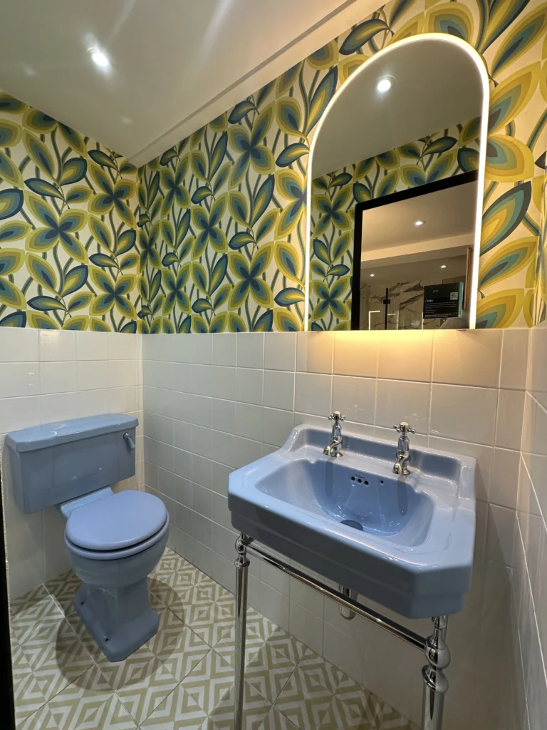 A bathroom with retro blue fixtures, floral wallpaper, geometric floor tiles, a lit mirror, and ambient ceiling lighting.