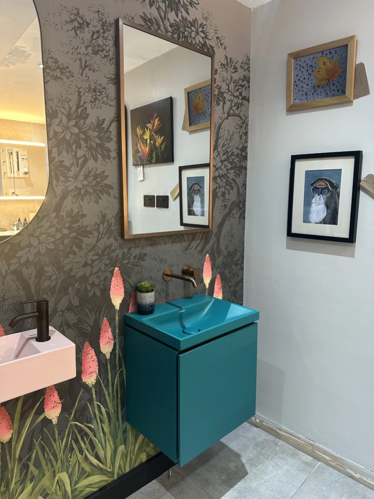 A stylish bathroom features floral wallpaper, teal cabinet, wall-mounted sink and faucet, and a mirror reflecting vibrant framed artwork.