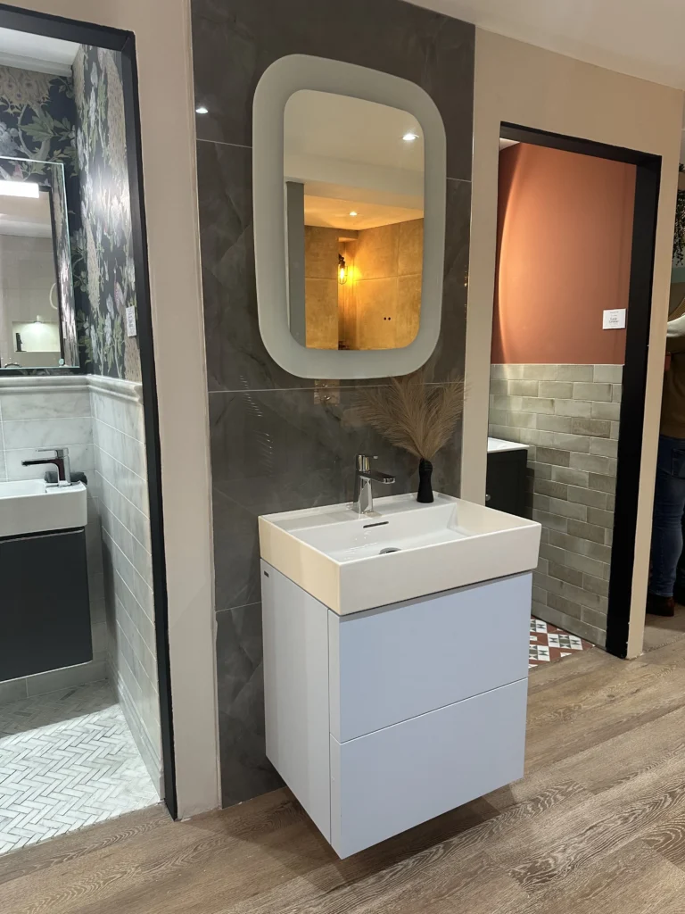 Modern bathroom display with sleek sink, large mirror, and patterned floors. Person stands partially visible in an adjacent room. Warm lighting highlights decor.