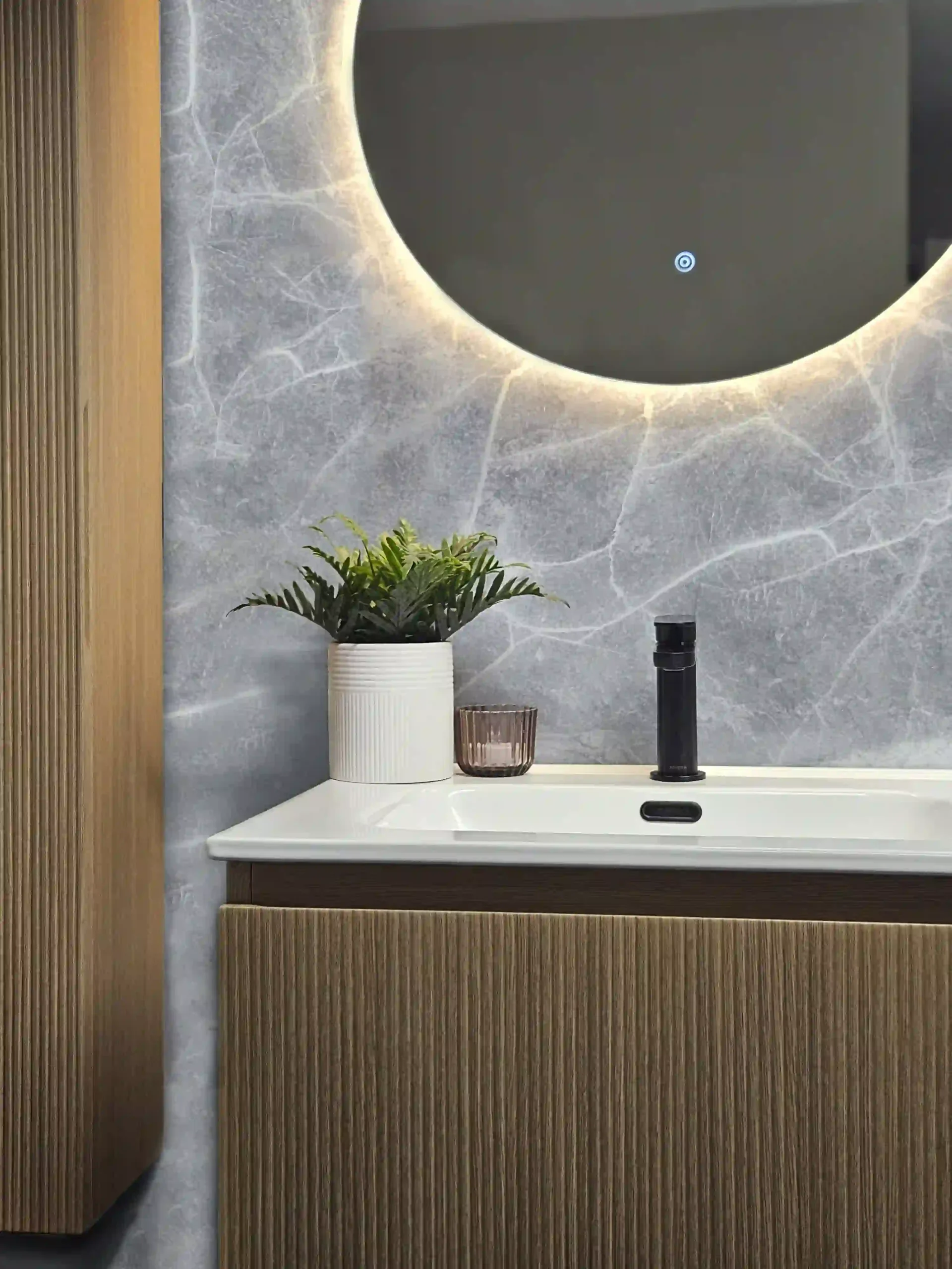 Modern bathroom vanity with backlit round mirror, potted plant, and textured wooden cabinetry. Elegantly designed with a minimalist aesthetic and subtle lighting.