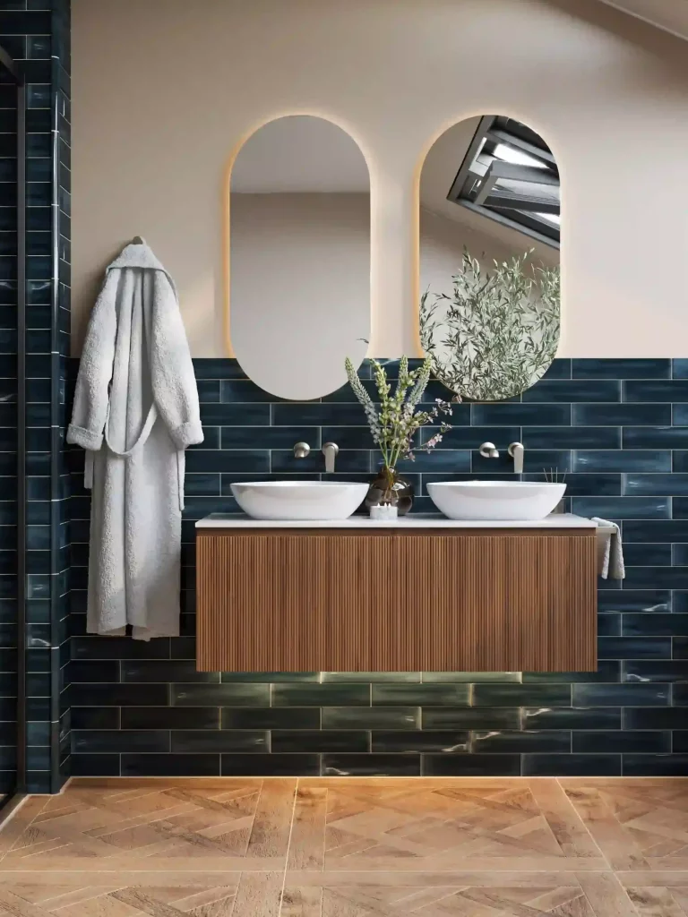 A modern bathroom features dual sinks, arch mirrors, green tiles, a wood vanity, and plants, with a robe hanging on the wall.